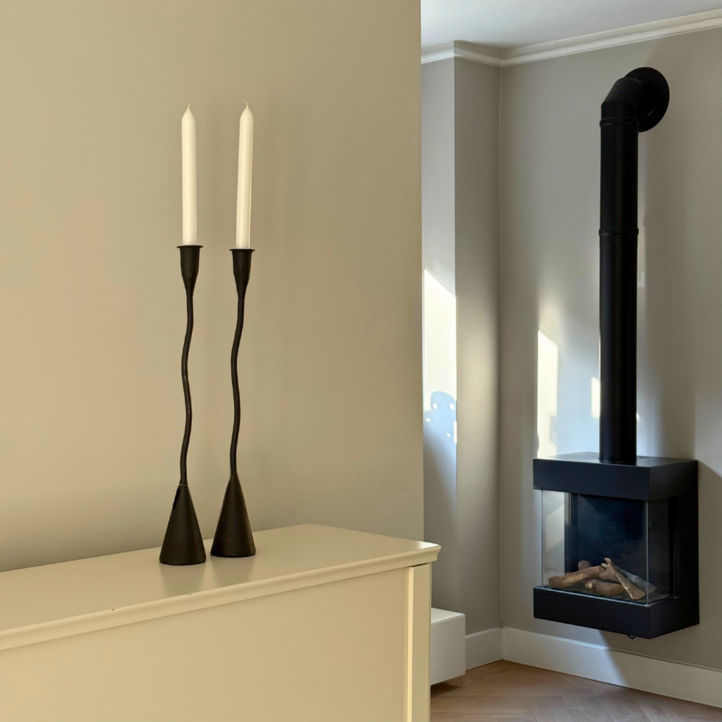 Twee golvende zwart metalen Askil kandelaars op een dressoir in een minimalistisch interieur met een gashaard