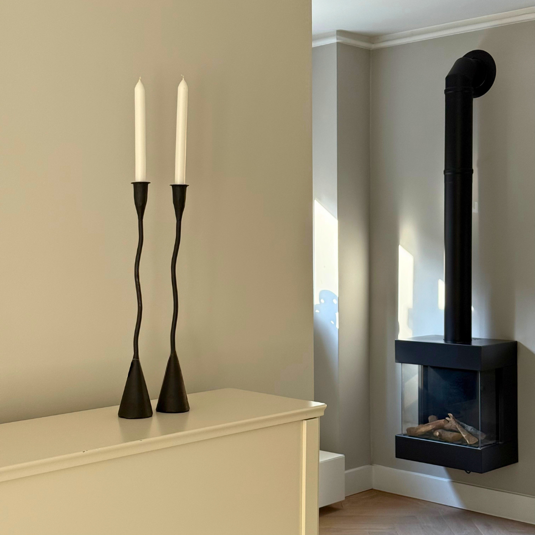 Twee golvende zwart metalen Askil kandelaars op een dressoir in een minimalistisch interieur met een gashaard