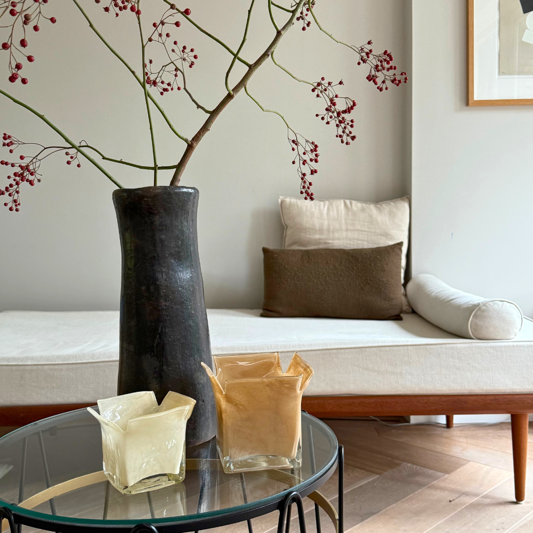 Zand- en vanillekleurige Atlas kaarshouders samen met een zwarte robuuste vaas op een glazen bijzettafel in een tijdloos interieur