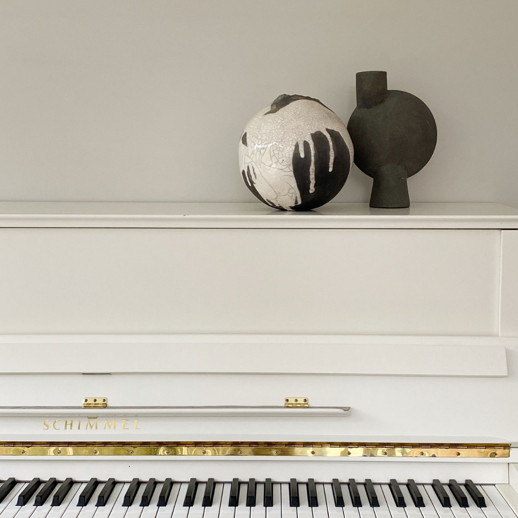 Bolvormige vaas van keramiek in creme en grijs tinten samen met een antraciet kleurige vaas op een witte piano