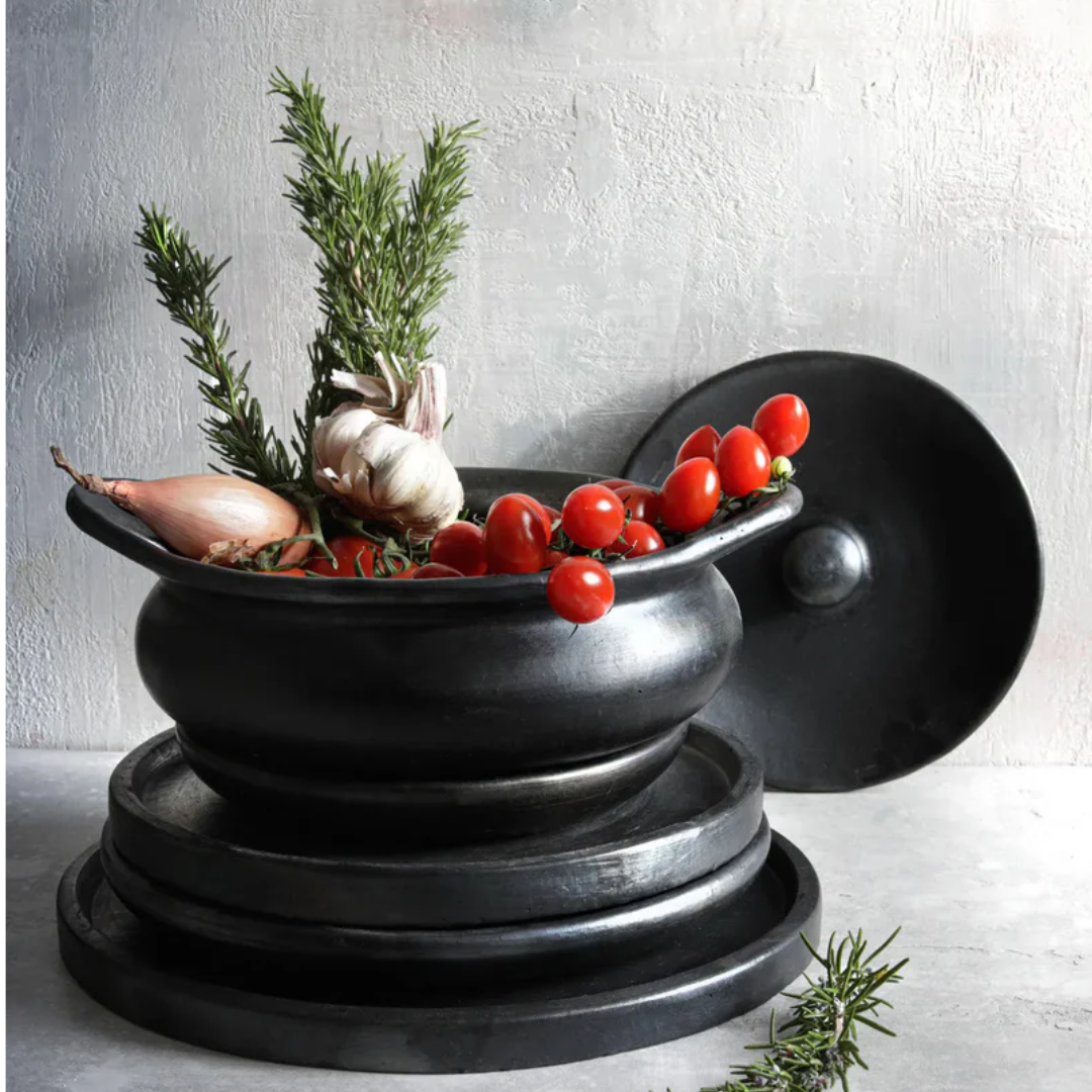 Stoere, minimalistische, handgemaakte pot van gebrand terracotta. Hier gevuld met tomaten, knoflook en rozemarijn bovenop een stapel borden uit de Burned collectie van Bazar Bizar.