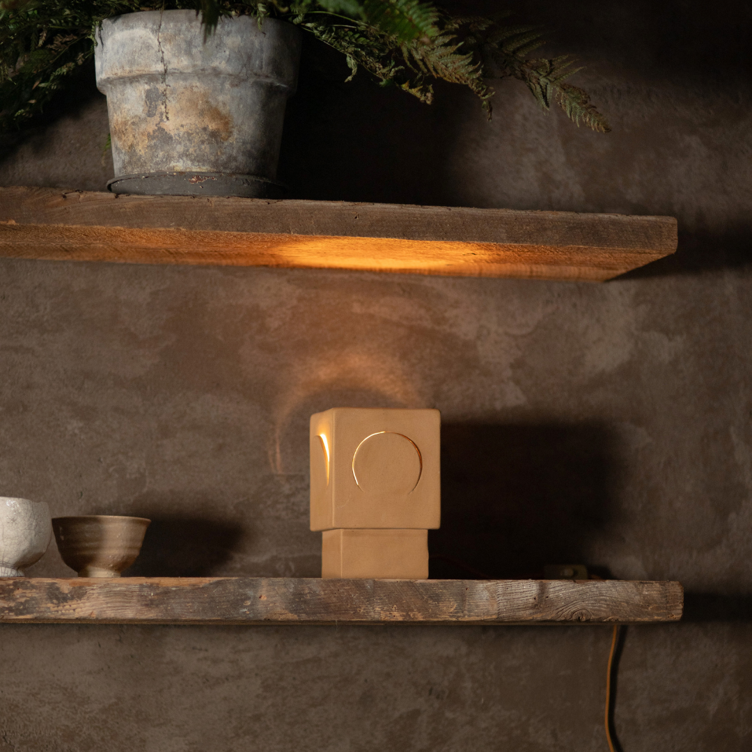 Kubistische tafellamp Eclipse van steengoed in oker op een houten wandplank in een warm interieur