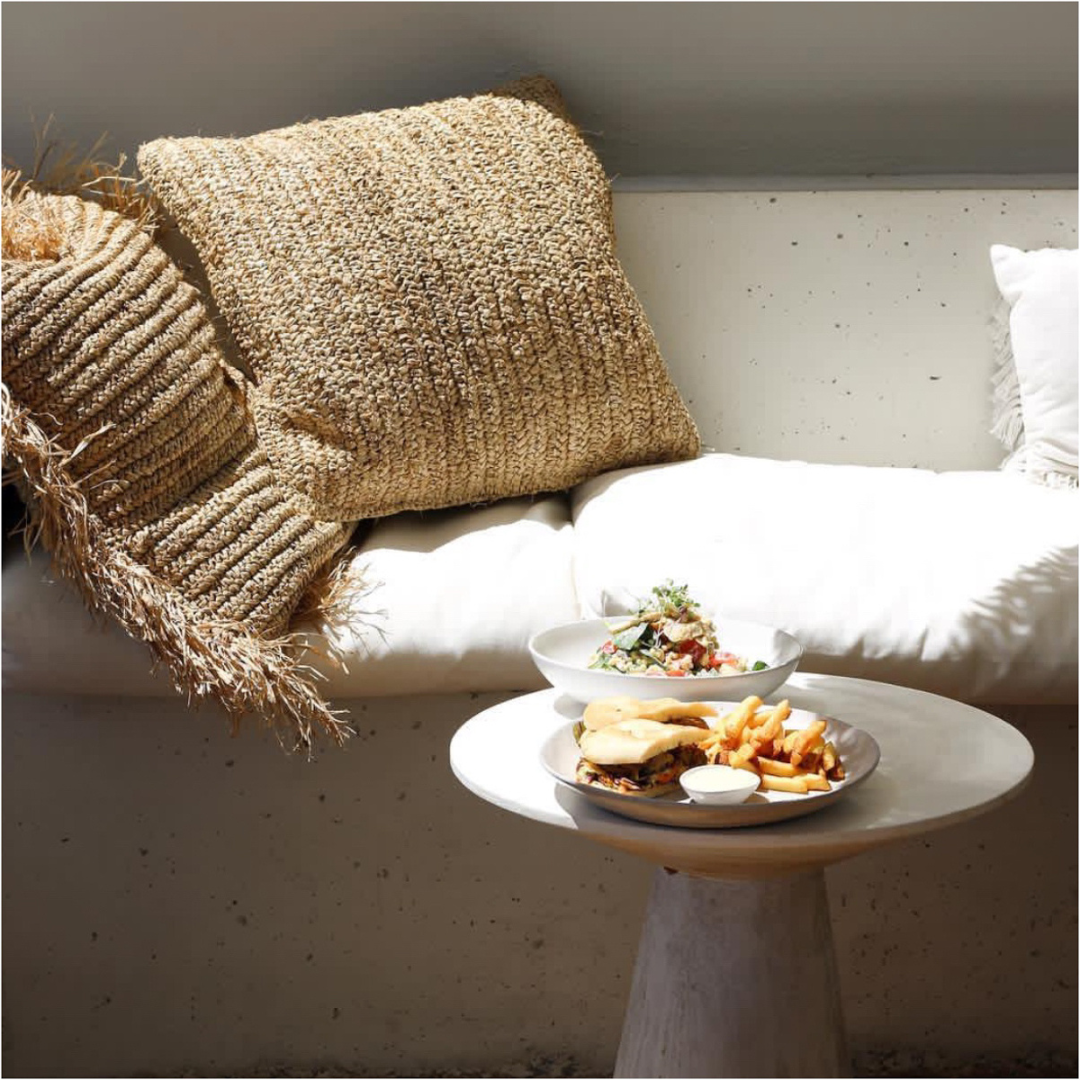 Raffia Flores kussenhoes op een witte loungebank naast een ronde betonnen bijzettafel met lunchgerechten