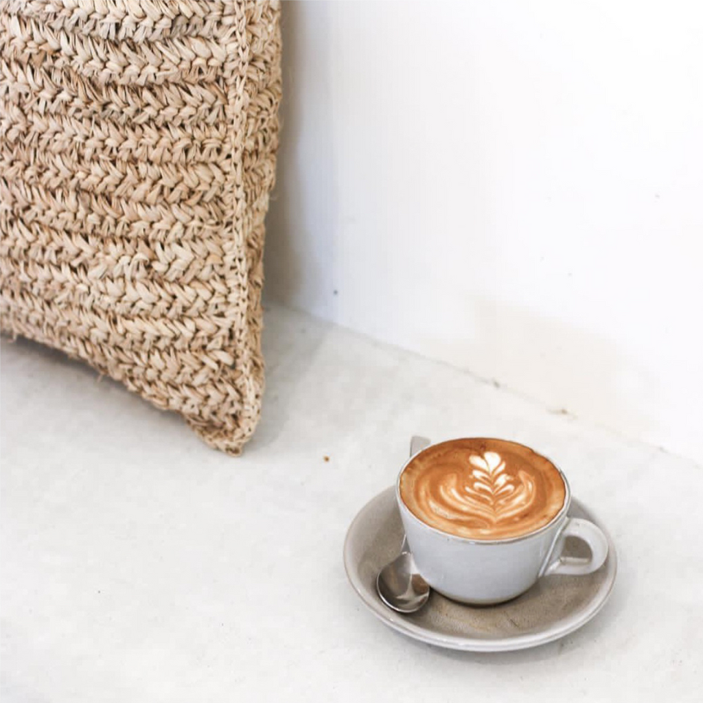 De Raffia Flores Kussenhoes van Bazar Bizar op een neutrale bank naast een kopje koffie